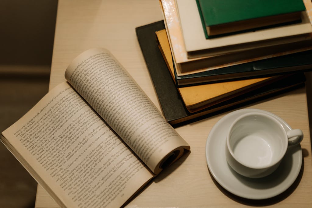 A cozy setup with open book, ceramic cup, and stacked books on a wooden table.