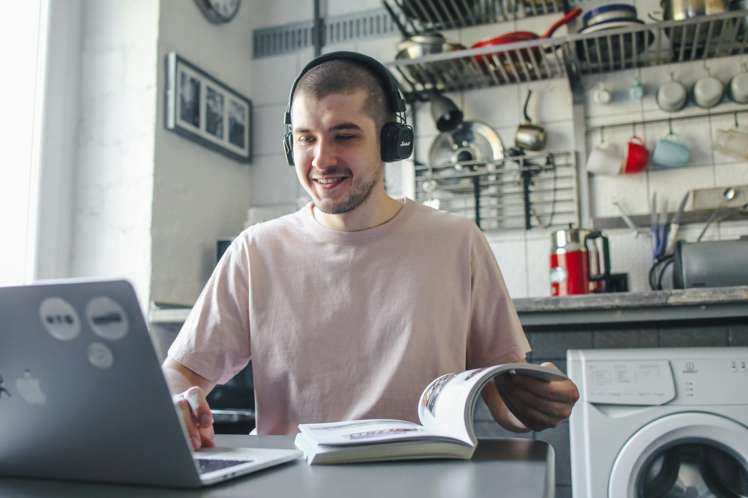 Smiling man wearing headphones, studying with a laptop and book in a cozy kitchen setting.
